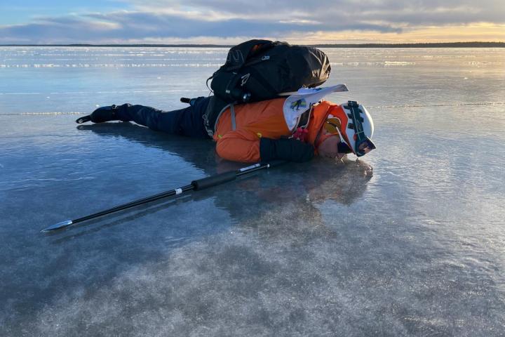 Person lying on ice with a backpack, wearing winter gear and using a pole for support.