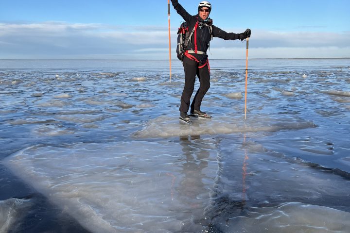 Person ice skating on a frozen lake, wearing winter gear and holding poles, with a clear blue sky.