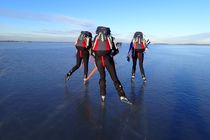 Three people ice skating on a vast frozen lake with backpacks under a clear blue sky.