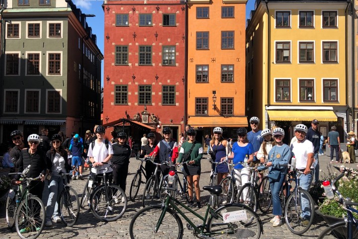 Group of cyclists in front of colorful buildings in a sunny plaza.