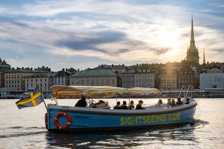 Sightseeing boat on water near city skyline at sunset with Swedish flag.