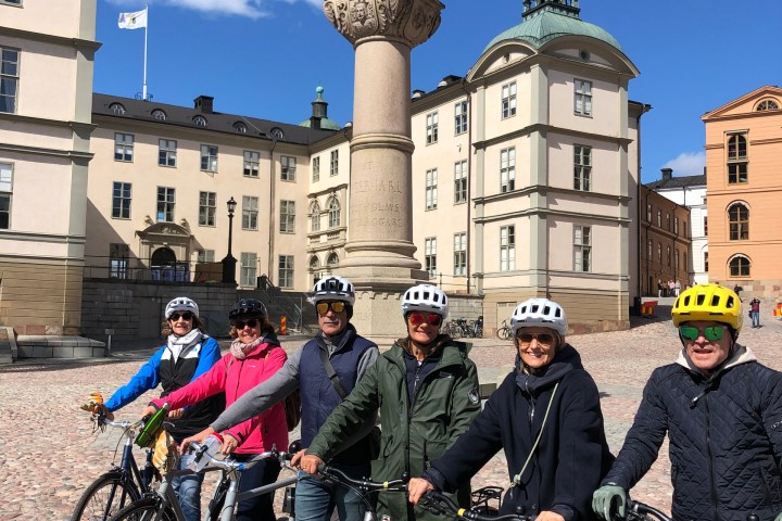 Group of six cyclists posing with bikes in front of a statue and historic buildings on a sunny day.