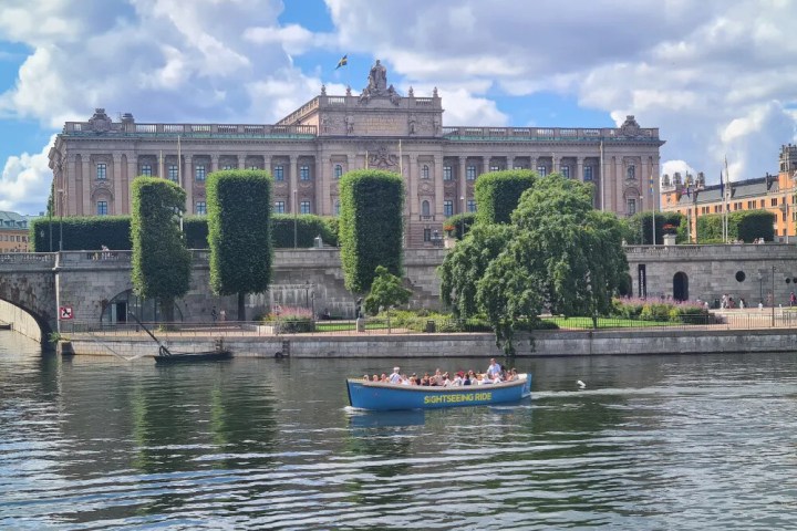 Boat on river with people, passing a historic building and manicured trees.